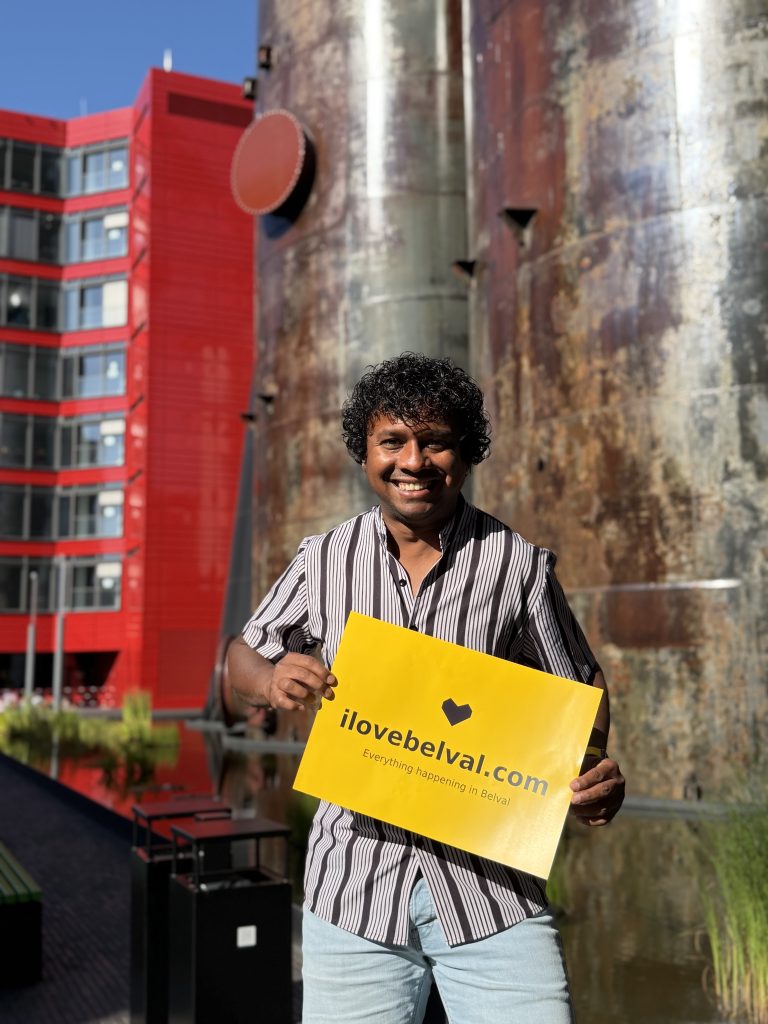 Aswin Lutchanah holding a yellow sign reading ilovebelval.com in front of the Belval blast furnaces, with the red Terres Rouges tower behind.
